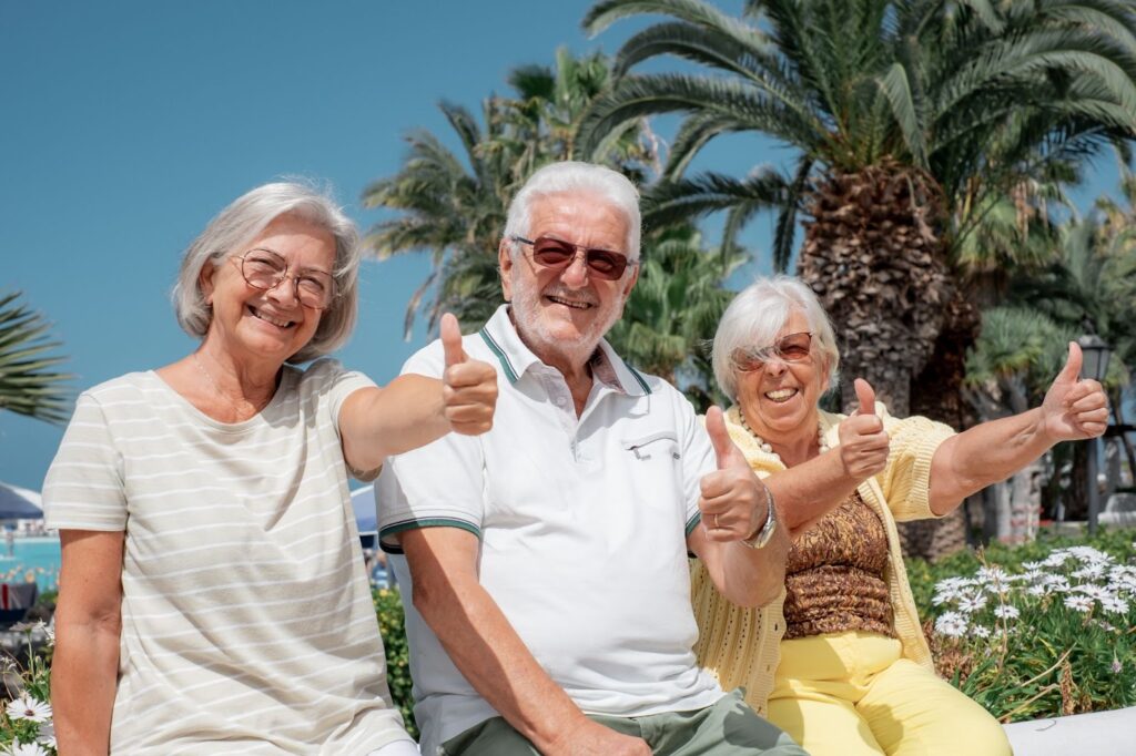 Three seniors smiling and giving a thumbs up as they sit in front of a large palm tree.