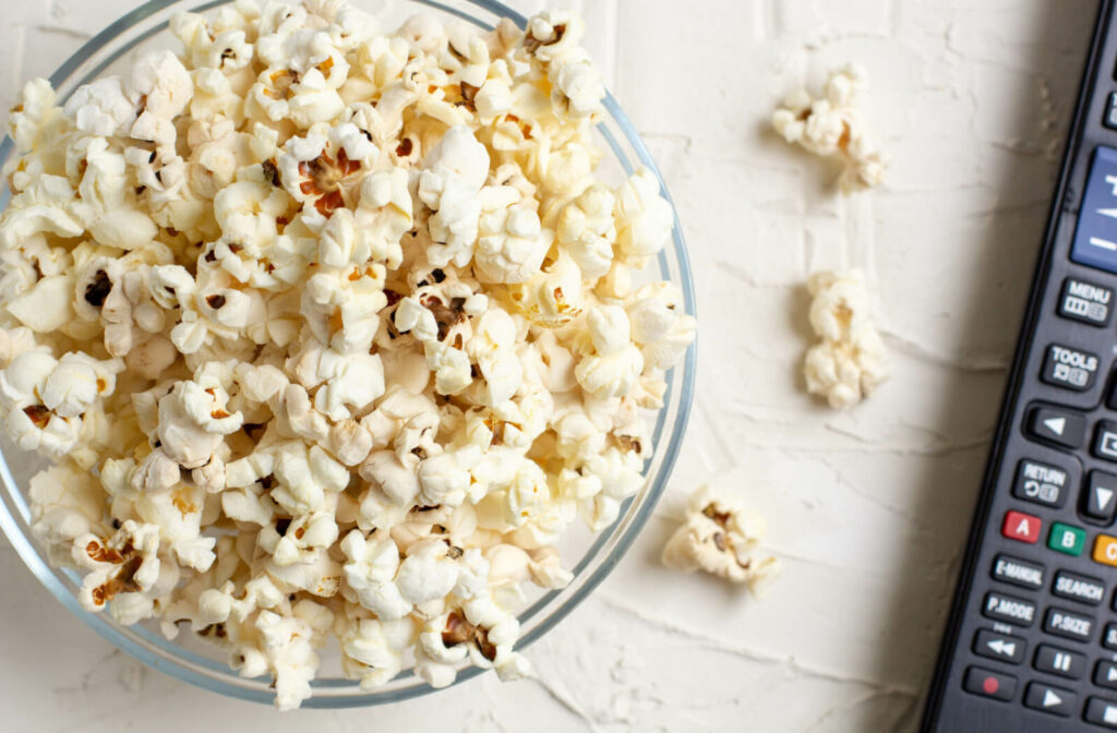 A bowl of popcorn in a clear bowl sitting on a white table beside a remote.