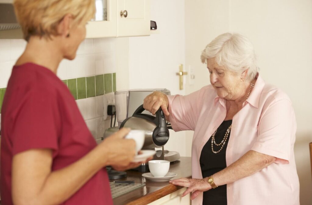 Senior woman pouring coffee into a cup in a home kitchen while a caregiver offers support.