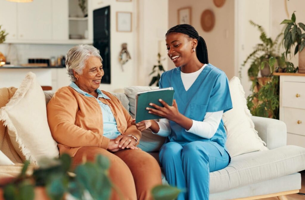 Caregiver sitting on a couch with an older adult while smiling and reading from a book or tablet in a cozy living space