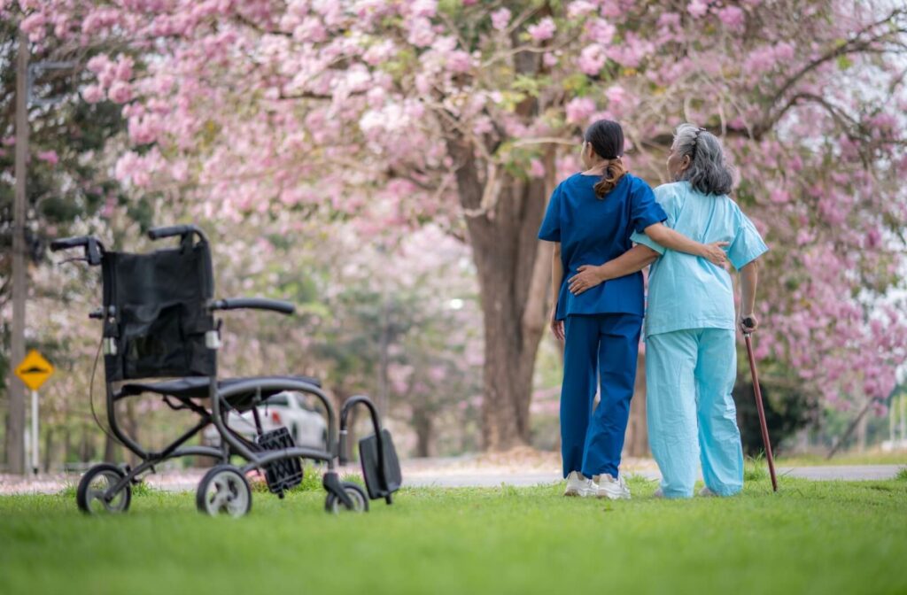 Caregiver walking arm-in-arm with an older adult outdoors near a wheelchair under blooming pink trees