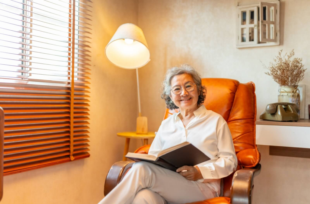 An older adult smiles while sitting in a comfortable leather chair under a lamp and reading a book in their living room.