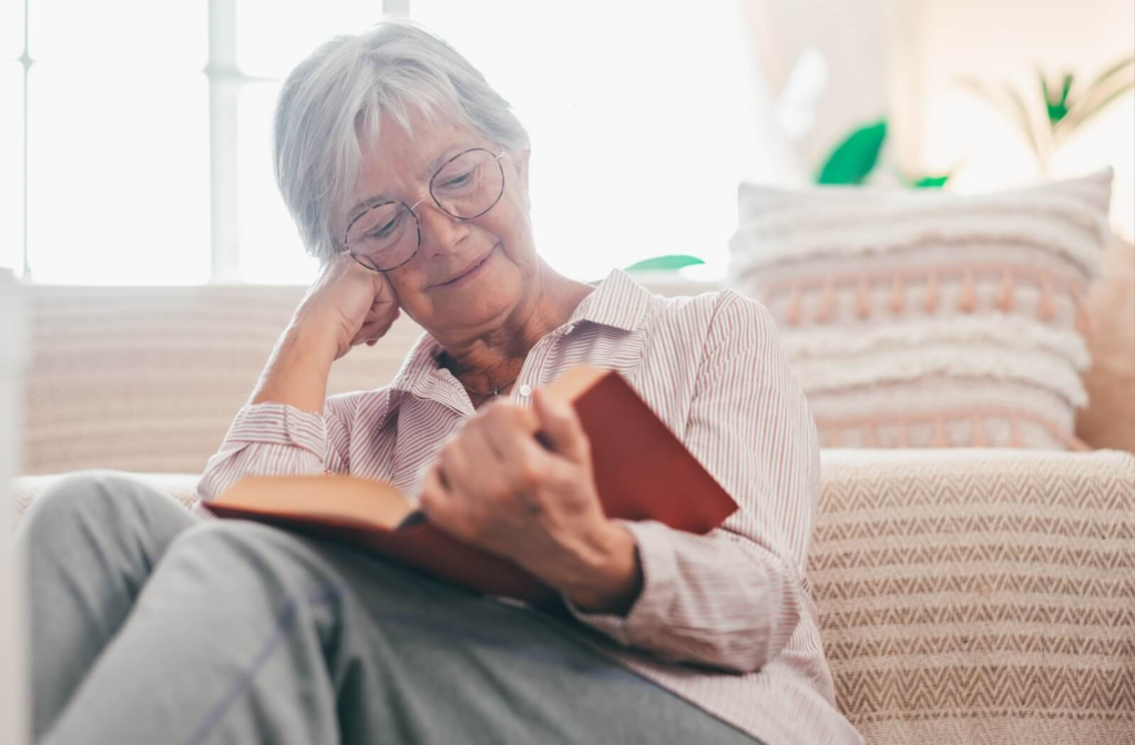 An older adult rests their face on their hand while reclining and reading a hardcover book on a pink couch covered in pillows.