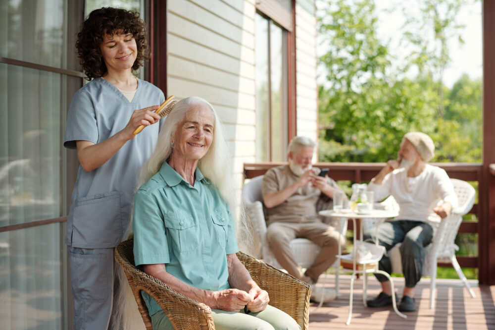 A happy young care partner brushing long white hair of smiling female resident of a memory care community on patio on summer morning against two aged men