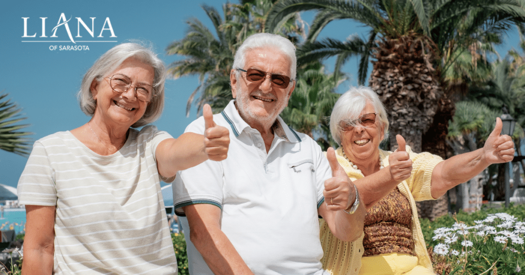 Three smiling senior citizens outdoors, giving thumbs up with palm trees and a clear blue sky in the background, enjoying an active and happy lifestyle.