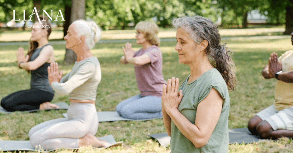 Group of older adults in a park performing balance exercises, kneeling on mats with hands pressed together in a meditation posture.