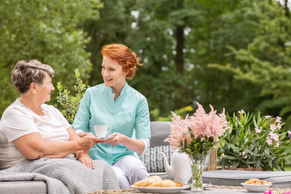 A smiling woman helping a senior woman with tea outside. Services and Amenities.