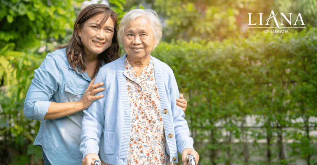 A care partneris supporting an older woman with a walker in the garden of a memory care community.